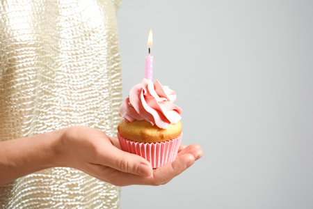 Woman holding delicious birthday cupcake with burning candle on light background, closeupの写真素材