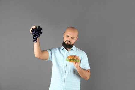 Overweight man with hamburger and grapes on gray background