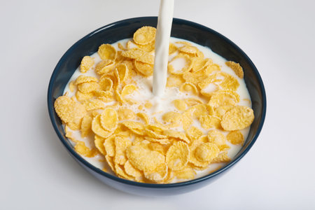 Milk pouring into bowl with crispy cornflakes on white background