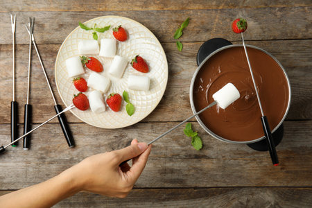 Woman dipping marshmallow into pot with chocolate fondue on wooden background, flat layの写真素材