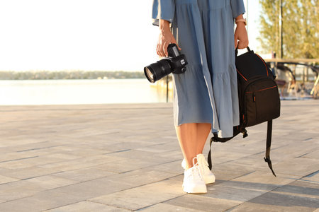 Young female photographer holding professional camera and backpack at pier. Space for textの写真素材