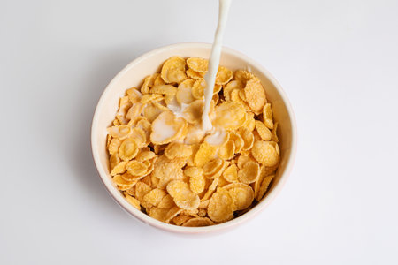 Milk pouring into bowl with crispy cornflakes on white background