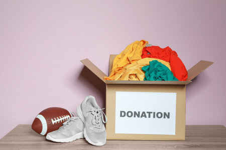 Donation box with clothes, shoes and rugby ball on table against color backgroundの写真素材