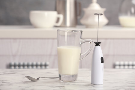 Milk frother device, glass cup and spoon on table in kitchenの写真素材
