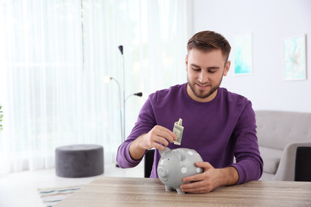 Young man putting money into piggy bank at table indoors. Space for textの写真素材
