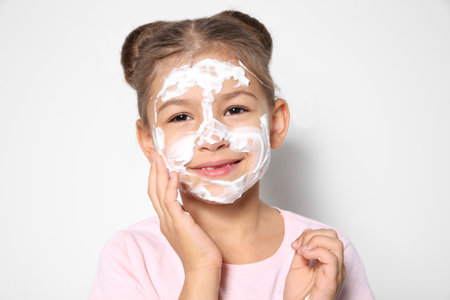 Cute little girl with soap foam on face against white backgroundの写真素材