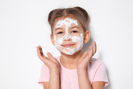 Cute little girl with soap foam on face against white backgroundの写真素材