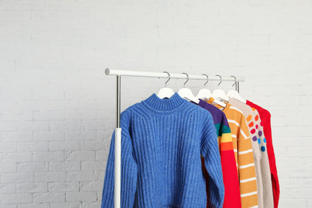 Collection of warm sweaters hanging on rack against white brick wallの写真素材