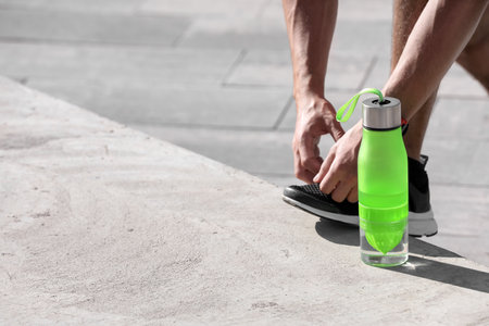 Sporty man tying shoelaces near bottle of water outdoors on sunny day. Space for textの写真素材