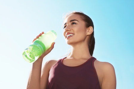 Young sporty woman drinking water from bottle against blue sky on sunny day