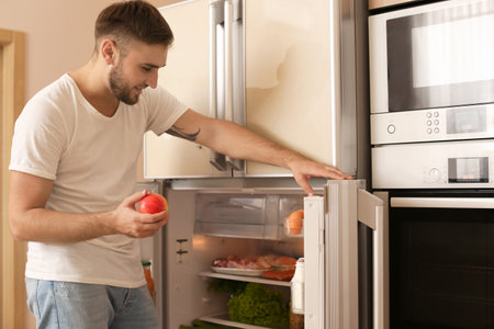 Young man taking apple from refrigerator in kitchenの写真素材