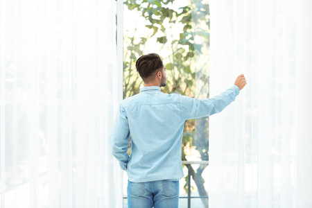 Young man opening window curtains at homeの写真素材