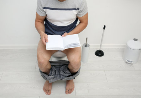Young man with book sitting on toilet bowl in bathroomの写真素材