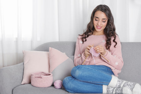 Young woman in warm sweater knitting on sofa at home. Space for textの写真素材
