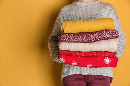 Woman holding stack of winter clothes on color background. Space for text