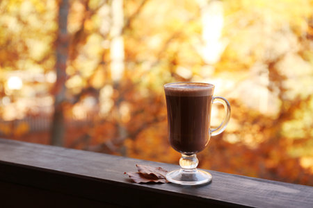 Hot cozy drink in glass cup and fallen autumn leaves on wooden railing at balcony. Space for textの写真素材