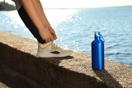 Young woman tying shoelaces near bottle of water at riverside on sunny dayの写真素材