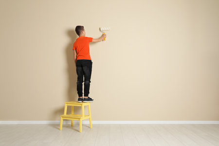 Child on step chair painting color wall with roller brush. Space for textの写真素材