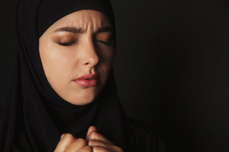 Portrait of sad Muslim woman in hijab praying on dark background. Space for textの写真素材
