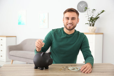 Young man putting coin into piggy bank at table indoorsの写真素材