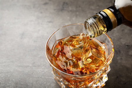 Pouring whiskey from bottle into glass with ice cubes on table. Space for textの写真素材