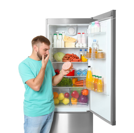 Young man with expired sausage near open refrigerator on white backgroundの写真素材