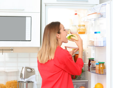 Hungry young woman eating sandwich near open refrigerator in kitchen. failed dietの写真素材