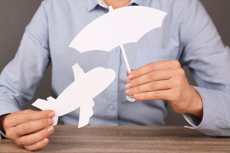 Insurance agent covering paper plane with umbrella cutout at table, closeup. Travel safety conceptの写真素材