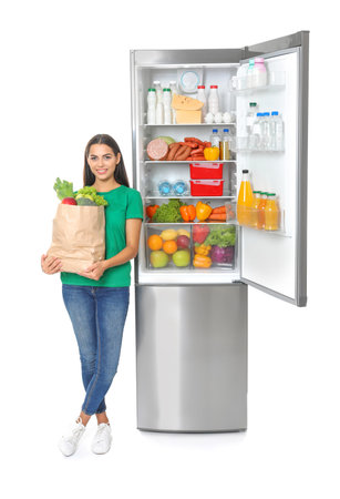 Young woman with bag of groceries near open refrigerator on white backgroundの写真素材