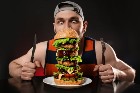 Young hungry man with cutlery eating huge burger on black backgroundの写真素材