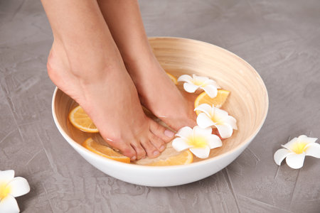 Woman soaking her feet in bowl with water, orange slices and flowers on gray background, closeup. spa treatmentの写真素材