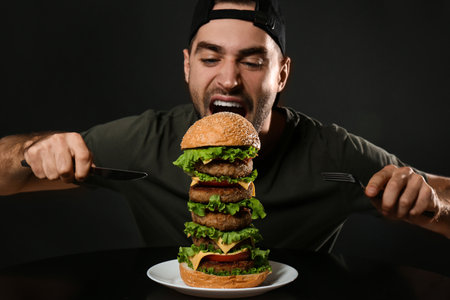 Young hungry man with cutlery eating huge burger on black backgroundの写真素材
