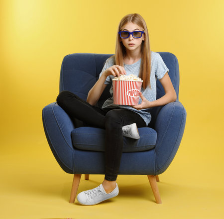 Emotional teenage girl with 3D glasses and popcorn sitting in armchair during cinema show on color backgroundの写真素材
