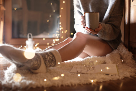 Woman with cup of hot beverage and book at home in winter evening, closeup