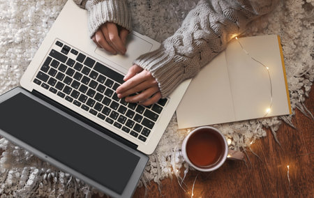 Woman with cup of hot beverage using laptop at home in winter evening, top view