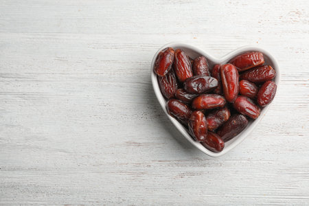 Heart shaped bowl with sweet dried date fruits on wooden background, top view. Space for textの写真素材