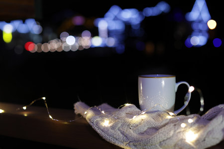 Cup of hot beverage, sweater and Christmas lights on wooden railing outdoors against blurred...