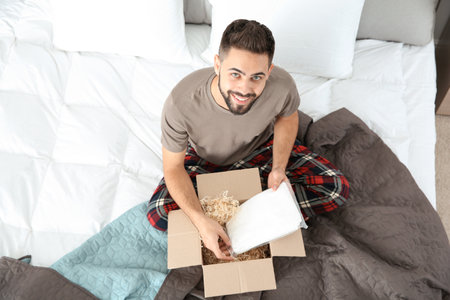 Young man opening parcel in bedroom at homeの写真素材