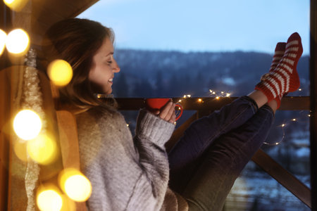 Woman with cup of hot beverage and Christmas lights resting on balcony. Winter eveningの写真素材