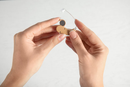 Woman putting battery into hearing aid on light background, closeup