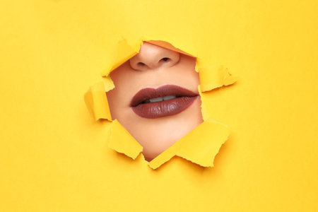 Lips of beautiful young woman with dark lipstick visible through hole in color paperの写真素材