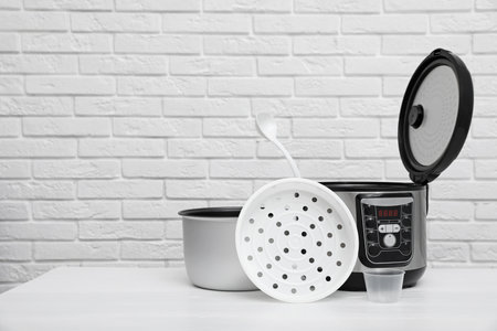 Modern electric multi cooker, parts and accessories on table near brick wall. Space for text