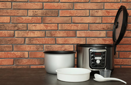 Modern electric multi cooker, parts and accessories on table near brick wall. Space for text