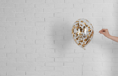 Young woman piercing bright balloon near brick wall, space for textの写真素材