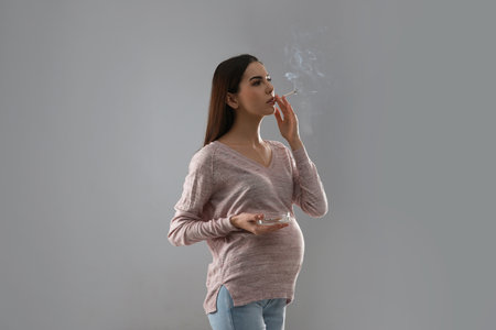 Young pregnant woman smoking cigarette on gray backgroundの写真素材