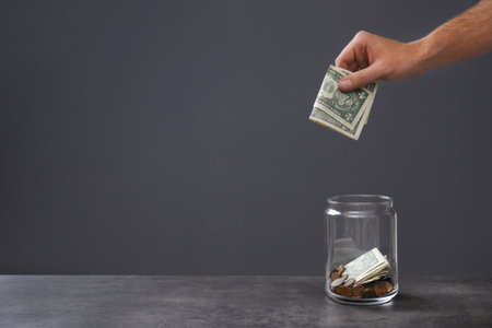 Man putting money into donation jar on table against gray background, closeup. Space for text