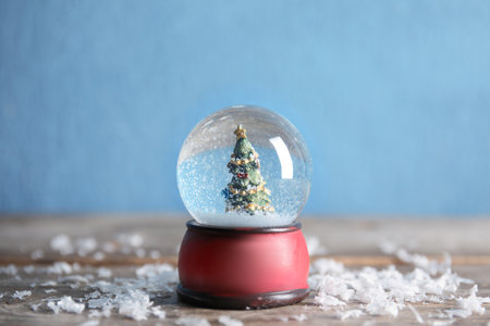 Magical snow globe with Christmas tree on wooden table against color background