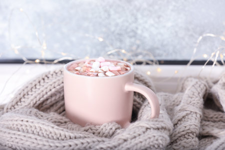 Cup of aromatic cacao with marshmallows and warm scarf on window sillの写真素材