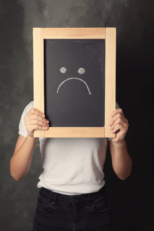 Woman hiding behind small chalkboard with drawing of sad face on dark background. depression symptomsの写真素材