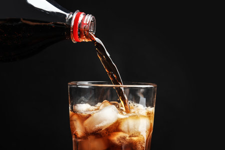 Pouring refreshing cola from bottle into glass with ice cubes on black background, closeupの写真素材
